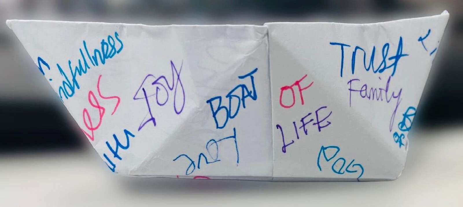 The original hand-folded paper boat that inspired Boat of Life Inc., covered with handwritten words including faithfulness, joy, trust, family, and love.